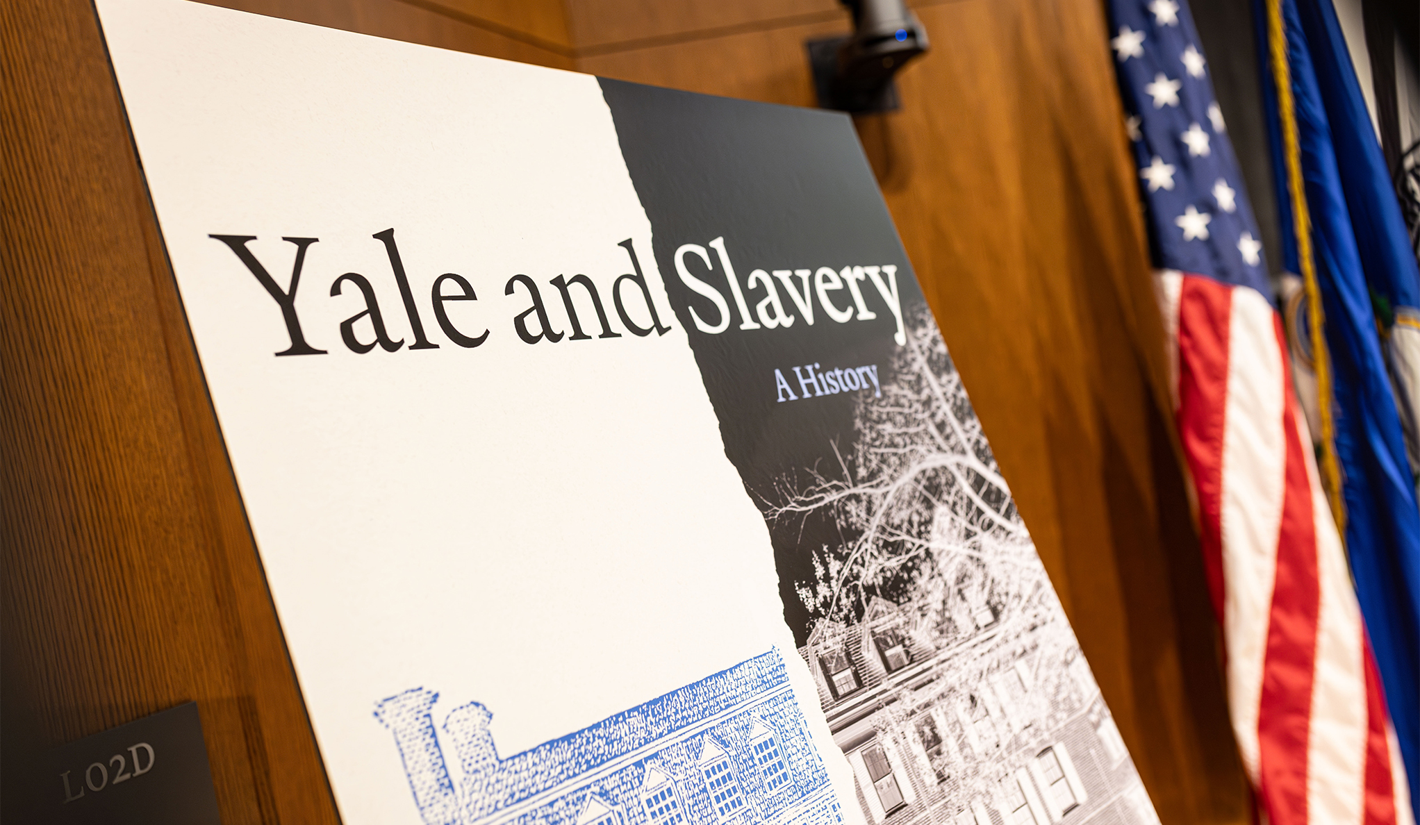 printed copy of the book cover for Yale and Slavery: A History on an easel in the front of a room with an American flag besides it.