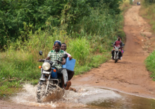 Two men ride a motorcycle through a puddle on a dirt road in Sierra Leone while holding a cooler full of vaccines.