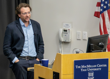 Peter Pomerantsev delivers a lecture at the MacMillan Center at Yale University while standing at a podium beside an American flag.