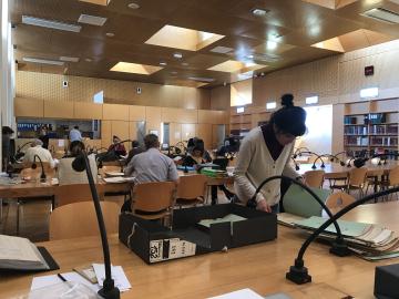 People work quietly at long wooden tables in a bright, modern reading room with high ceilings and natural light. Several individuals are examining archival documents and folders.