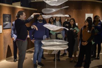 A group of visitors listens as a man gestures toward a hanging layered sculpture made of translucent material during a gallery tour. The group stands attentively around the installation, which is illuminated in a warmly lit exhibition space.
