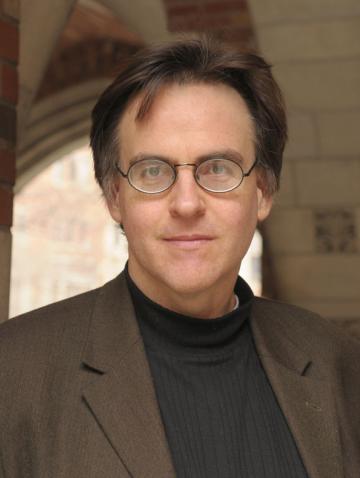 John MacKay under a brick archway wearing a black shirt, brown suit, and round glasses