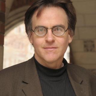 John MacKay under a brick archway wearing a black shirt, brown suit, and round glasses
