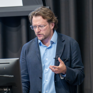 Peter Pomerantsev speaker delivers a lecture at the MacMillan Center at Yale University, gesturing with his hands while standing at a podium beside an American flag.