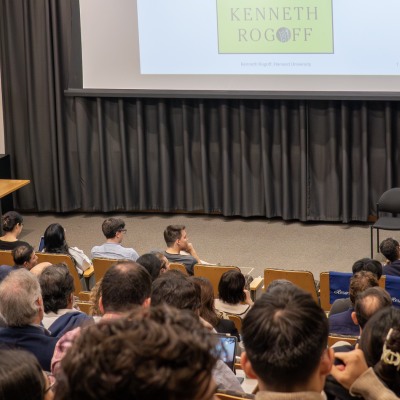 Kenneth Rogoff stands at a wooden podium bearing the Yale University MacMillan Center logo, delivering remarks in a lecture setting. The individual wears a dark blue suit and light blue shirt and appears engaged while speaking into a microphone. An American flag is visible in the background.