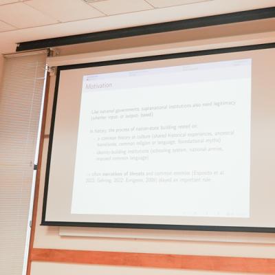 A visiting scholar in a red sweater presents with hands expressed in front of a projector.